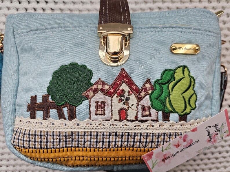 May include: A light blue canvas handbag with a brown leather handle and gold clasp. The bag features a patchwork design with embroidered houses, trees, and a fence. The bag has a white lace trim and a yellow and brown plaid fabric lining. The bag has a tag that says "Ybbs & Creativity Handmade with love".