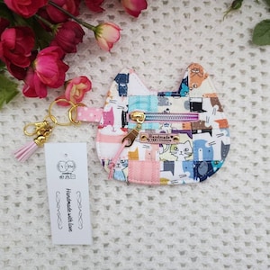 May include: A pink and white cat-shaped coin purse with a zipper closure and a gold key chain. The purse is made of fabric with a patchwork design featuring various colorful cats. The tag attached to the key chain reads "Handmade with love".