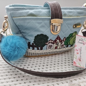 May include: A light blue quilted canvas handbag with a brown strap, a gold clasp, and a small blue pom-pom keychain. The bag features an embroidered design of houses and trees. The strap has a tag that reads "Yoli's Creations Handmade with Love".