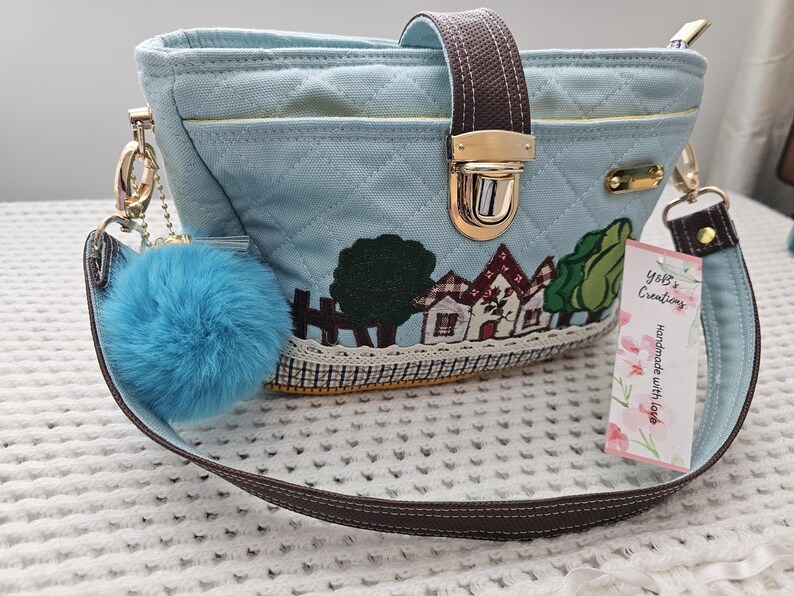 May include: A light blue quilted canvas handbag with a brown strap, a gold clasp, and a small blue pom-pom keychain. The bag features an embroidered design of houses and trees. The strap has a tag that reads "Yoli's Creations Handmade with Love".
