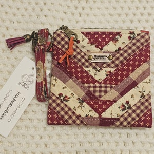 May include: A small red and white patchwork fabric zippered pouch with a floral print and a matching fabric wrist strap with a tassel. The pouch has a metal zipper pull and a small metal tag that reads "Handmade by Tiff's Creations".