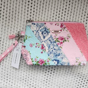May include: A pink and blue patchwork fabric zippered pouch with a pink and white polka dot wrist strap. The pouch has a floral print and the strap has a tassel. The pouch has a label that says "Handmade with love".