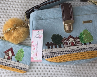 May include: A light blue quilted handbag and matching coin purse featuring embroidered house and tree designs, white lace trim, and yellow fabric borders. A brown strap with a gold metal clasp and a fluffy mustard yellow pom-pom charm are attached.