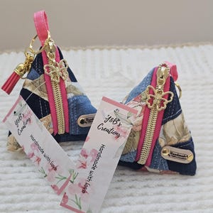 May include: Two handmade triangle-shaped coin purses. They feature a patchwork design with denim and floral fabrics, accented with pink zippers and butterfly charms. Each purse has a pink strap and a tag that reads "y&b's Creations, Handmade with love."