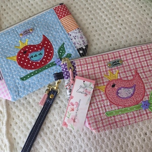 May include: Two quilted zipper pouches with wrist straps, one light blue and one pink, both featuring a red or pink bird appliqué with a yellow crown, green leaves, and a purple flower. Each pouch has a Yob's Creations label and a hanging tag.