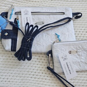 May include: Two white quilted fabric purses with blue denim accents and a blue and white strap. The larger purse has a zipper closure and the smaller purse has a snap closure. Both purses have a tag that says "Handmade with love".