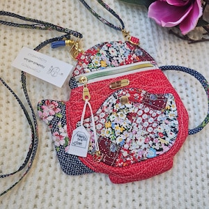 May include: A red and floral patterned teapot-shaped crossbody bag with a black and white polka dot handle. The bag has a zipper closure and a small pocket on the front. The bag is made of fabric and has a gold chain strap.