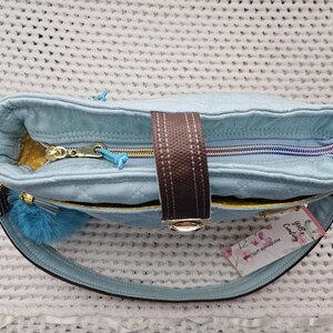 May include: A light blue quilted tote bag with a brown strap and a gold zipper. The bag has a fluffy blue pom-pom charm attached to the strap and a tag that says "Lily's Candy".
