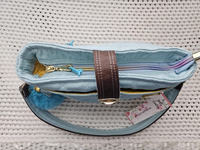 May include: A light blue quilted tote bag with a brown strap and a gold zipper. The bag has a fluffy blue pom-pom charm attached to the strap and a tag that says "Lily's Candy".