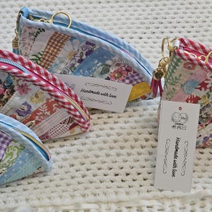 May include: Three small, triangular fabric pouches with zipper closures and keychains. The pouches are made of patchwork fabric in shades of blue, pink, and white with floral and geometric patterns. Each pouch has a tag that reads "Handmade with love".