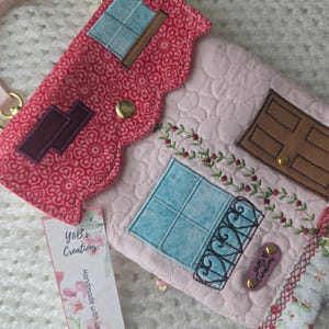 May include: A pink quilted house-shaped crossbody bag with a red patterned roof and floral accents. Features a brown door, blue windows, and a pink strap. Includes a tag that says "YoB's Creations".