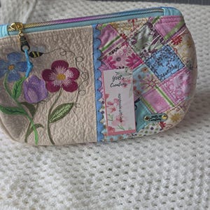 May include: A quilted zipper pouch featuring a beige side with embroidered blue, purple, and magenta flowers and a bee, and a patchwork side with floral patterns in pink, blue, and white. A tag reads Y&B Creations Handmade with Love.