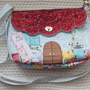 May include: A handmade, quilted crossbody bag with a house design. The bag features a red roof, a brown door, and a light blue body with floral and patchwork details. The bag has a light blue strap and a tag that says "Yob's Creations".