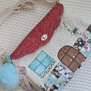 May include: A handmade quilted crossbody bag with a red flap and a light blue pom-pom. The bag features a patchwork design with a window and door applique. It has a cream-colored adjustable strap and gold-colored hardware.