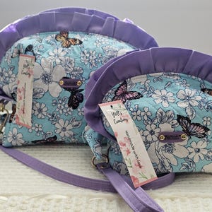 May include: Two matching handbags with a floral and butterfly pattern. The bags have a light blue background with white flowers and butterfly accents. The trim and straps are purple. Each bag has a tag that reads "YoB's Creations".