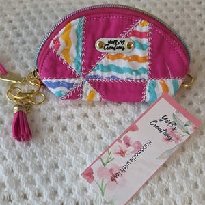 May include: A semi-circular coin purse featuring a vibrant pink fabric with a multi-colored striped patchwork pattern. It includes a gold-tone zipper, a matching pink tassel keychain, and a metal tag labeled Y&B's Creations. A rectangular tag is attached.