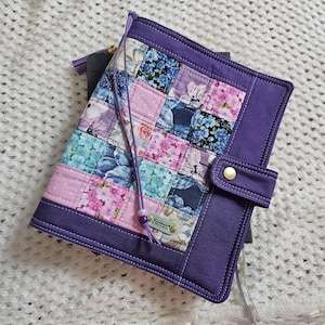 May include: Purple fabric notebook cover with a patchwork design of floral and geometric patterns. The cover has a flap closure with a gold button and a purple strap.