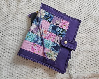 May include: A quilted fabric notebook cover featuring a patchwork design of floral and polka dot patterns in shades of purple, blue, and pink. The cover has a solid purple border, a snap button closure, and a purple cord with a small bead and tag.