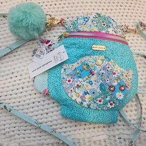 May include: A small turquoise blue quilted bag with a floral print and a zipper closure. The bag has a matching turquoise blue strap with white polka dots and a fluffy turquoise blue pom-pom keychain. The bag has a small pocket on the front and a tag that says "Handmade with love".