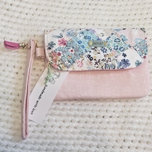 May include: A pink fabric clutch with a floral print and a flap closure. The clutch has a pink strap with a tassel and a tag that says "Handmade with love".