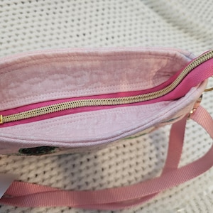 May include: A pink fabric bag with a gold zipper and a pink strap. The bag has a floral pattern.
