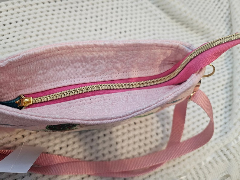 May include: A pink fabric bag with a gold zipper and a pink strap. The bag has a floral pattern.