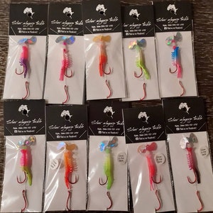 May include: Twelve individually packaged fishing lures with holographic accents. Each lure features a shrimp design with a variety of colors, including pink, green, blue, and red. Some lures have a glow-in-the-dark feature. The packaging includes the brand name "Silver Alyers Tackle" and the company's website.