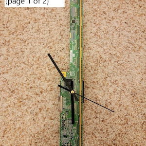 Op de afbeelding: Een groene printplaat met gouden connectoren is gemonteerd op een klok. De wijzers van de klok zijn zwart en wijzen naar 12 en 3. De printplaat is gelabeld met "G - Circuit Board (page 1 of 2)".