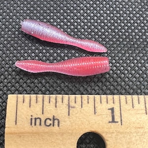 Puede incluir: Dos señuelos de pesca de plástico blando en color rosa translúcido con partículas de purpurina fina. Los señuelos presentan un diseño de cuerpo segmentado y acanalado con una cola cónica. Una regla de madera muestra que miden 2,5 cm.