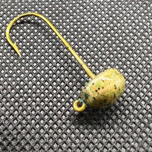 May include: A gold-colored fishing hook with a green and black speckled lure. The hook is curved with a sharp point, and the lure is oval-shaped. The hook and lure are set against a dark, textured background.