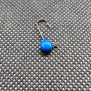 Könnte beinhalten: Ein einzelner blauer Jigkopf mit einem goldfarbenen Haken. Der runde Kopf hat eine leuchtend blaue Farbe. Der Haken ist gebogen und hat eine scharfe Spitze. Der Jigkopf ist zum Angeln bestimmt.