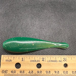 Puede incluir: Un señuelo de pesca de plástico blando verde con un cuerpo delgado y alargado. El señuelo mide aproximadamente 7,6 cm de largo y tiene una superficie texturizada con pequeñas motas de color. Una regla de madera está debajo.