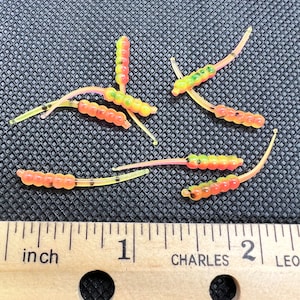 May include: A close-up of a ruler measuring a group of 7 small, flexible, yellow, orange, and pink fishing lures. The lures are about 2 inches long and have a segmented body.