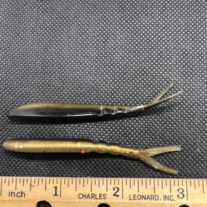 May include: Two soft plastic fishing lures with a split tail design. The top lure is black and bronze, while the bottom lure is bronze with gold and red glitter. A wooden ruler is positioned below, showing the lures are approximately 3 inches long.