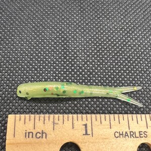 May include: A translucent green fishing lure with glitter accents. The lure is approximately 1 inch long and has a forked tail. The word "inch" is visible on a ruler next to the lure.