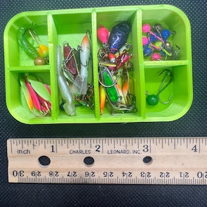 Puede incluir: Una caja de aparejos de plástico verde brillante llena de varios señuelos de pesca. Los señuelos incluyen spinners coloridos, jigs y cebos de plástico blando. Una regla con medidas en pulgadas está en primer plano.