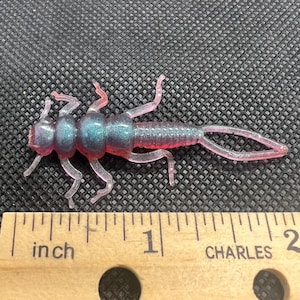 May include: A translucent pink and blue fishing lure shaped like an insect. The lure has multiple legs and a segmented body. The lure is approximately 1 inch long.