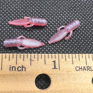 Puede incluir: Tres señuelos de pesca pequeños de color rosa translúcido con detalles de purpurina, cada uno con forma de crustáceo con cuerpo segmentado y apéndices finos. Una regla de madera se sitúa bajo los señuelos, mostrando que miden unos 2,5 cm.