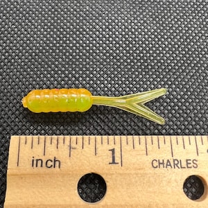 Peut inclure: Un petit leurre de pêche jaune et orange avec un corps segmenté et une queue fourchue. Le leurre mesure environ 2,5 cm de long, comme l'indique la règle sur l'image. Le leurre est conçu pour attirer les poissons.