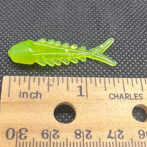 Puede incluir: Un señuelo de pesca translúcido de color verde neón con forma de pez pequeño, con costados acanalados y cola bifurcada, colocado sobre una regla de madera que muestra una longitud de unos 5,7 centímetros desde la cabeza hasta la cola.