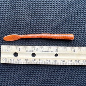 Op de afbeelding: Een bruine, gesegmenteerde, wormvormige vislokaas, ongeveer 7,6 cm lang, met een gevlekte, afgeronde kop. Het lokaas ligt op een zwart, getextureerd oppervlak naast een liniaal.