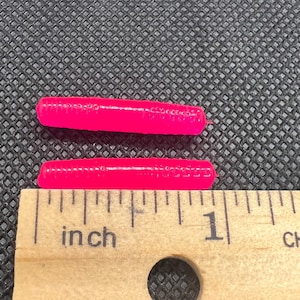 Puede incluir: Dos señuelos de pesca cilíndricos de color rosa brillante con textura acanalada, colocados horizontalmente sobre una regla de madera. Los señuelos miden aproximadamente 3,8 cm de largo, según las marcas en la herramienta de medición.