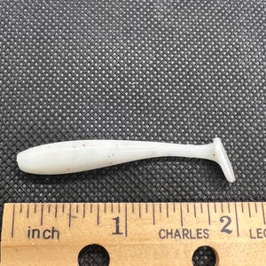 May include: A white soft plastic fishing lure with a paddle tail, speckled with red and blue dots. The lure is approximately 2 inches long and lies next to a wooden ruler.