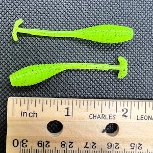 Puede incluir: Dos señuelos de pesca de plástico blando de color verde lima con una superficie texturizada. Los señuelos miden aproximadamente 5 cm de largo y tienen un anzuelo en un extremo.