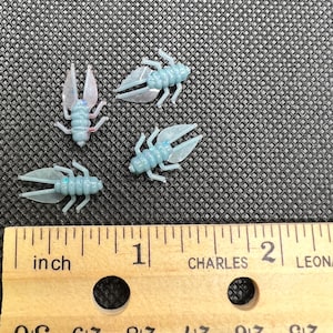 Puede incluir: Cuatro pequeños amuletos con forma de insecto azul claro con alas transparentes y detalles plateados. Una regla de madera con medidas en pulgadas está en primer plano. Los amuletos están sobre una superficie negra texturizada.