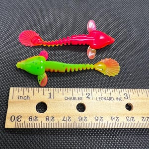 Puede incluir: Dos señuelos de pesca de plástico, uno rojo y otro verde, con un cuerpo segmentado y una cola en forma de abanico. Los señuelos se miden con una regla, mostrando que tienen aproximadamente 5 cm de largo.