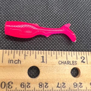 Könnte beinhalten: Ein leuchtend neonpinker Angelköder aus weichem Kunststoff in Form eines kleinen Fisches mit Paddelschwanz. Der Köder liegt über einem Holzlineal und zeigt eine Länge von etwa 5 Zentimetern vom Kopf bis zur Schwanzspitze.