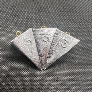 May include: Three dark gray fishing weights with a triangular shape and a gold-colored loop at the top. Each weight has a number embossed on its surface. The weights are arranged on a textured black surface.