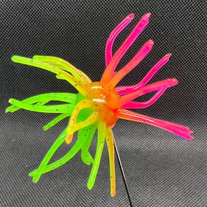 Könnte beinhalten: Ein farbenfroher Angelköder mit einem orangefarbenen Mittelteil und mehreren neon-grünen und rosa Tentakeln. Die Tentakel haben kleine schwarze Sprenkel. Der Köder soll Fische anlocken und ist an einem dünnen Metalldraht befestigt.