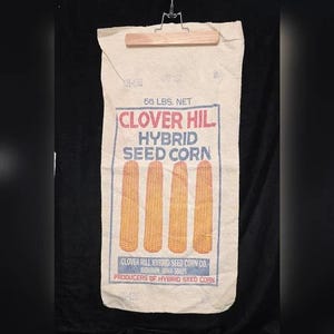 May include: A vintage Clover Hil Hybrid Seed Corn bag. The bag is cream-colored with a wooden clamp at the top. The bag features the text "CLOVER HIL HYBRID SEED CORN" in red and blue, along with illustrations of four ears of corn. The bag weighs 56 lbs.
